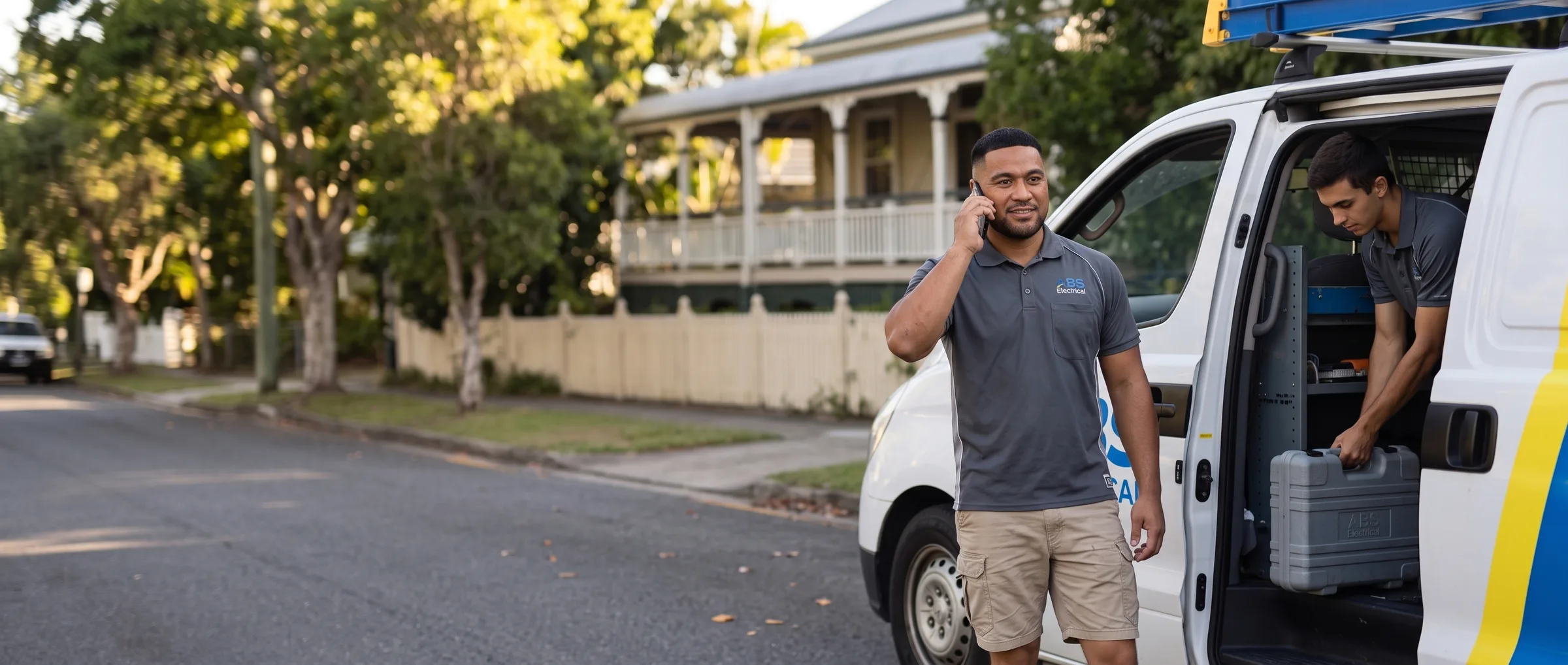 ABS electrician on the phone beside the van while an apprentice unloads tools